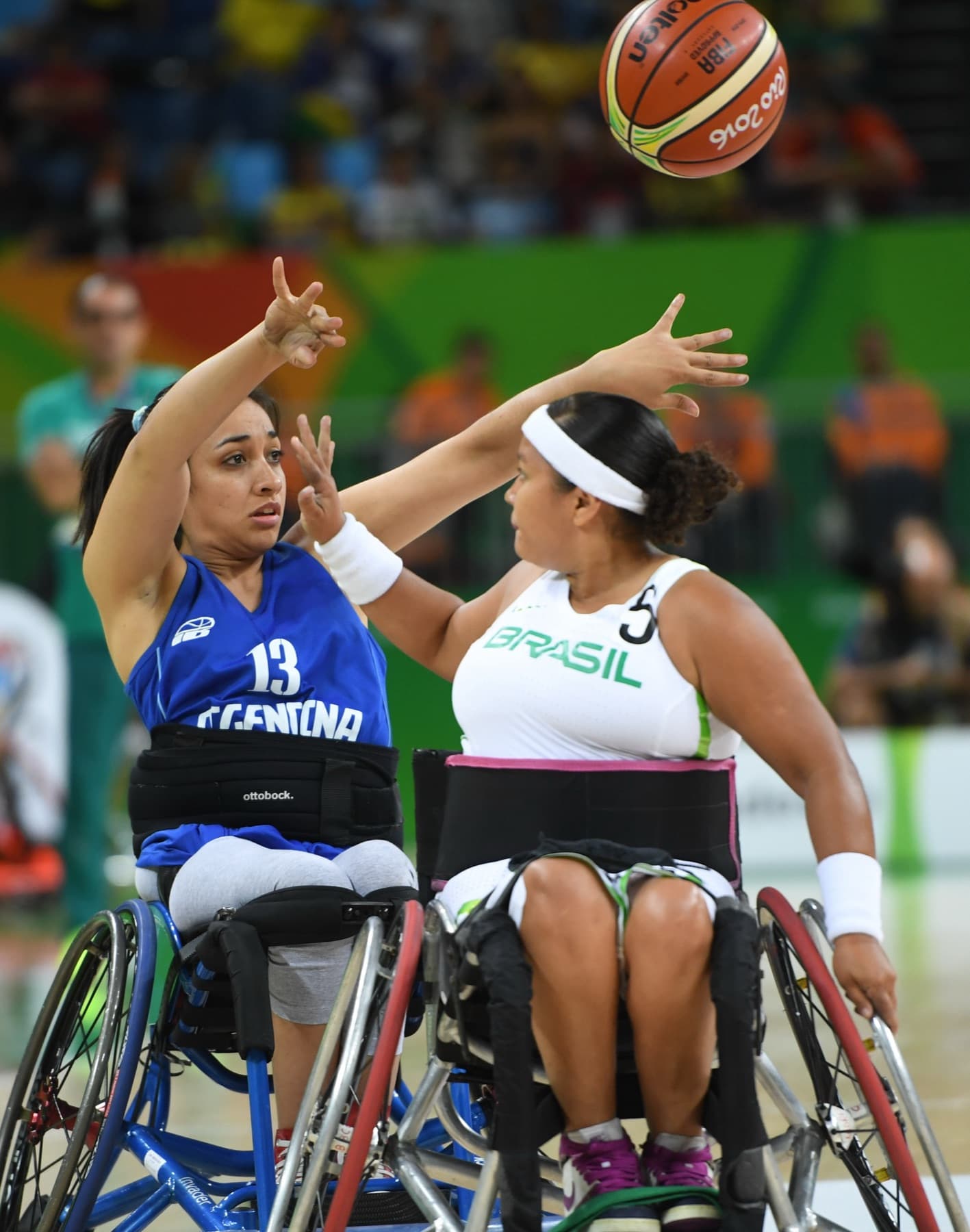 Jogadoras em cadeira de rodas esticam os braços na disputa pela bola