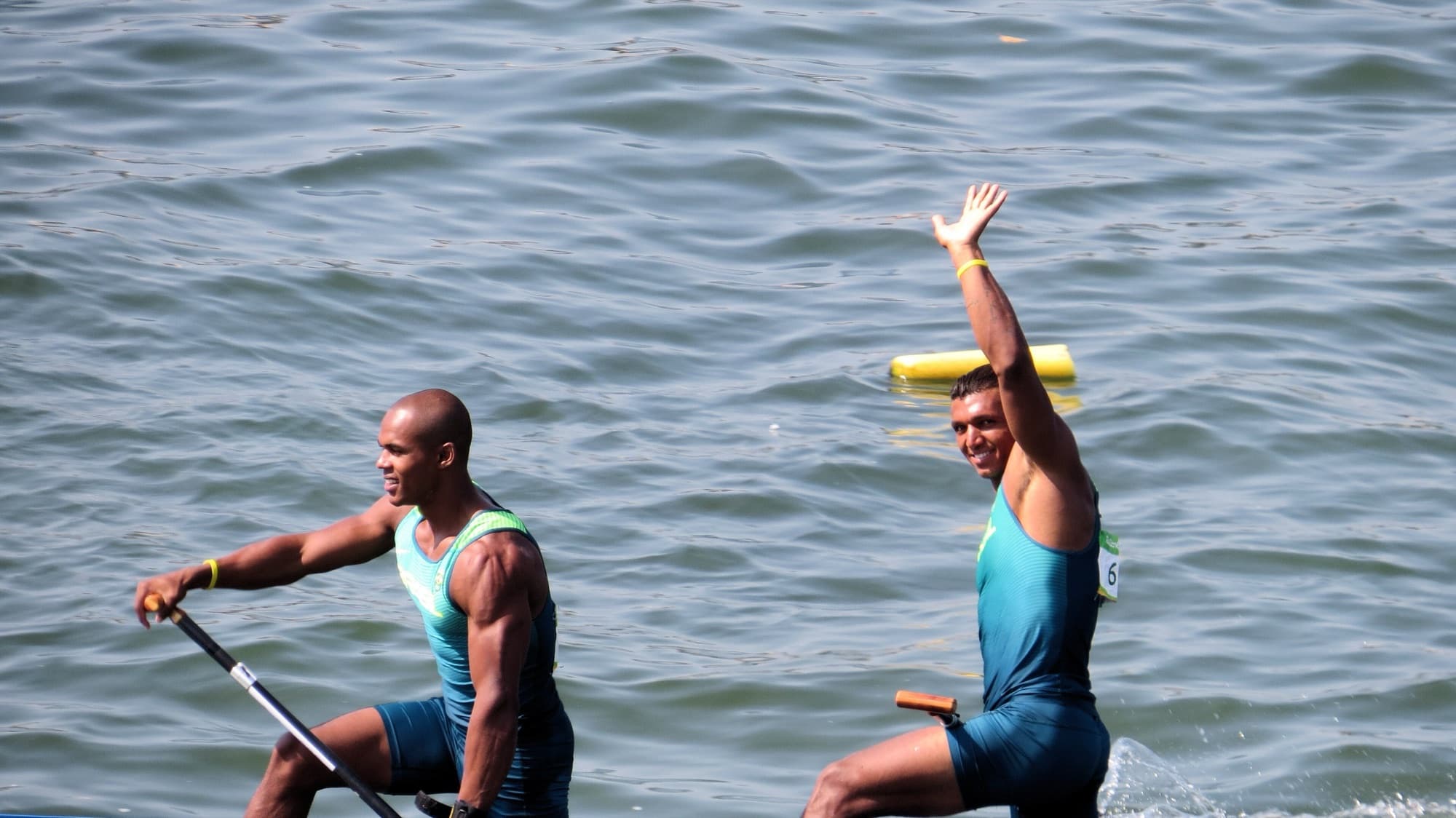 Dois canoístas brasileiros remam lado a lado; um deles acena para a arquibancada