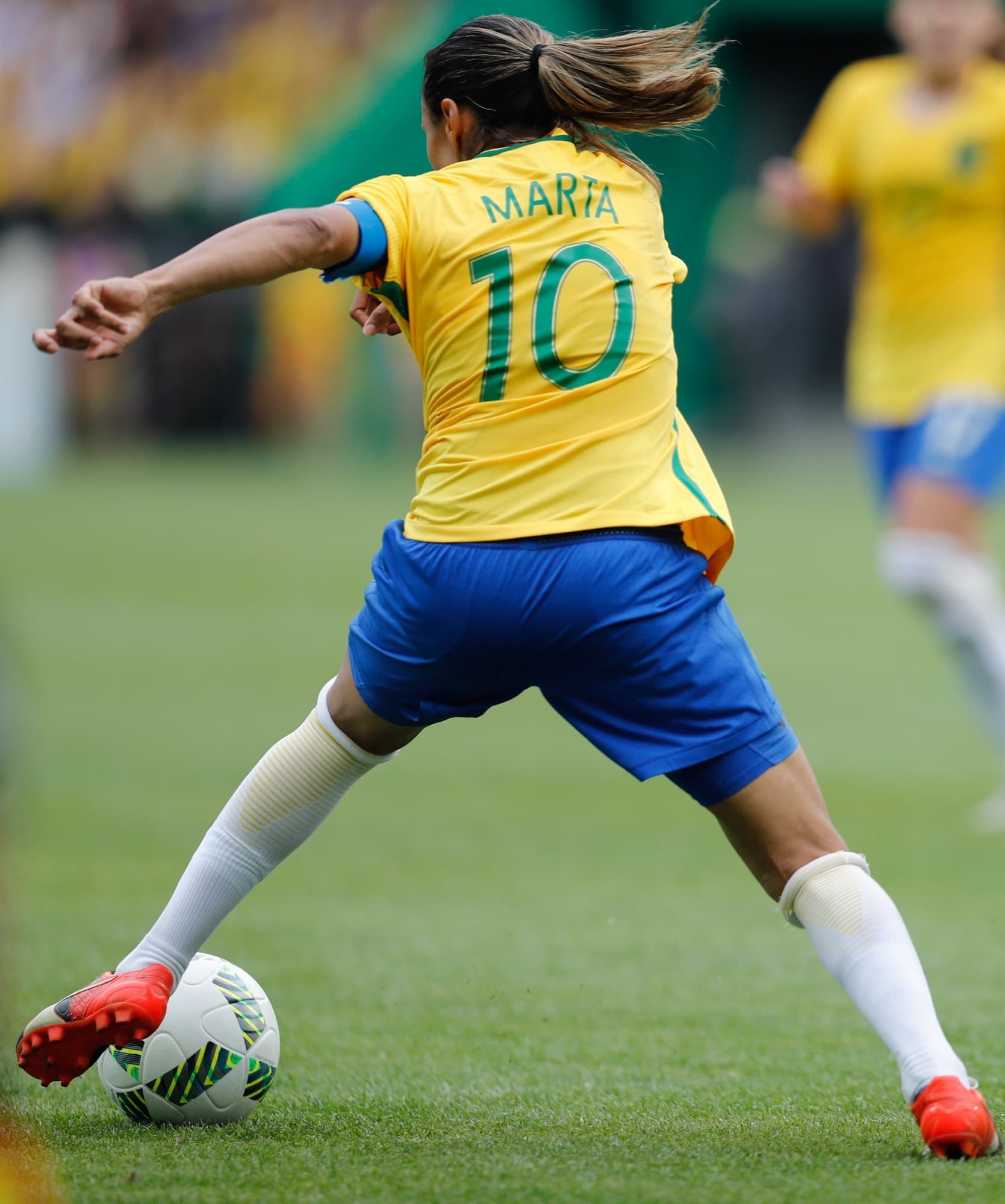 Jogadora da seleção brasileira, de costas, arma o chute com a bola no pé esquerdo no gramado