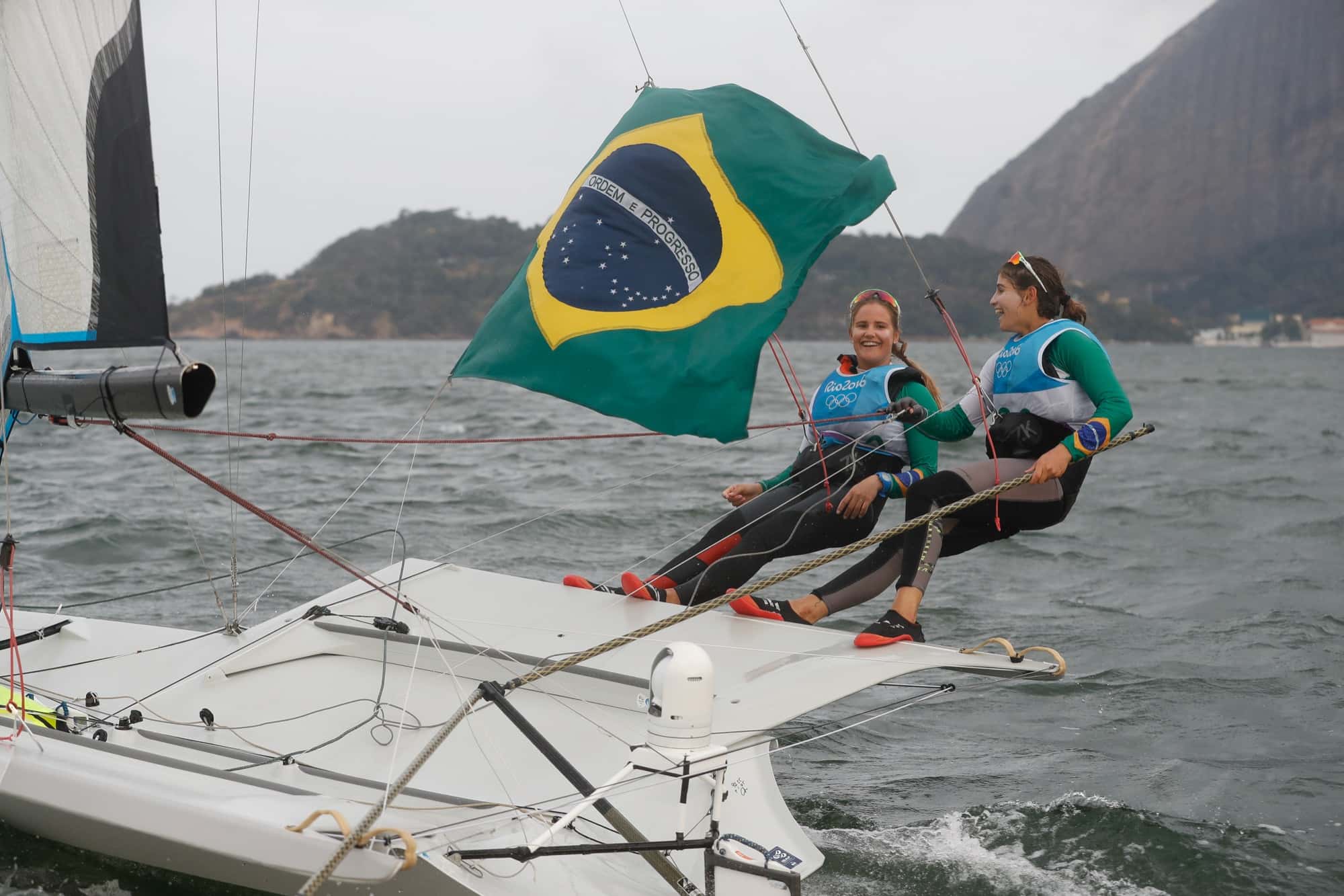 Dupla de velejadoras navega no barco com a bandeira do Brasil tremulando sobre elas
