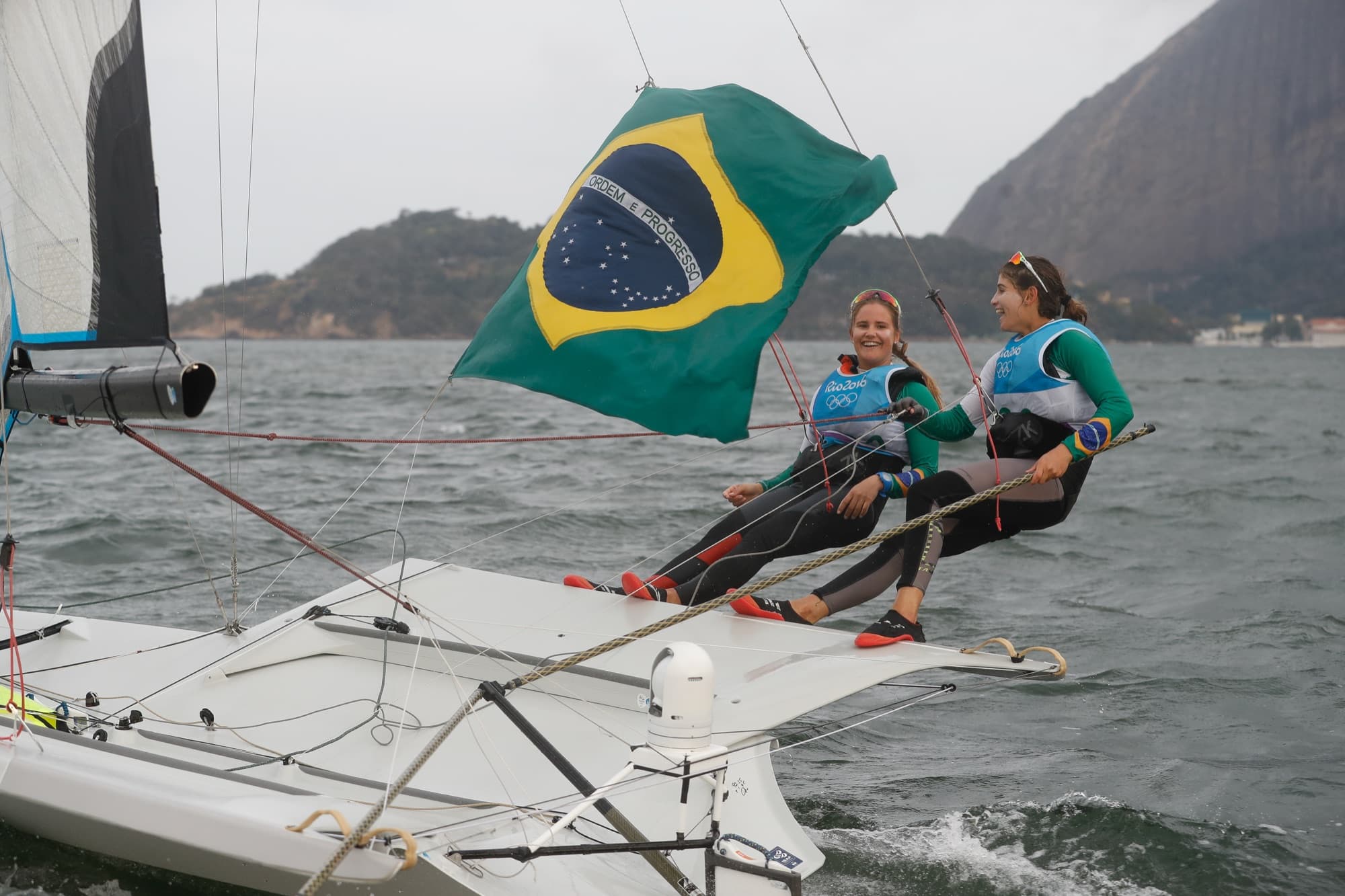 Dupla de velejadoras navega no barco com a bandeira do Brasil tremulando sobre elas