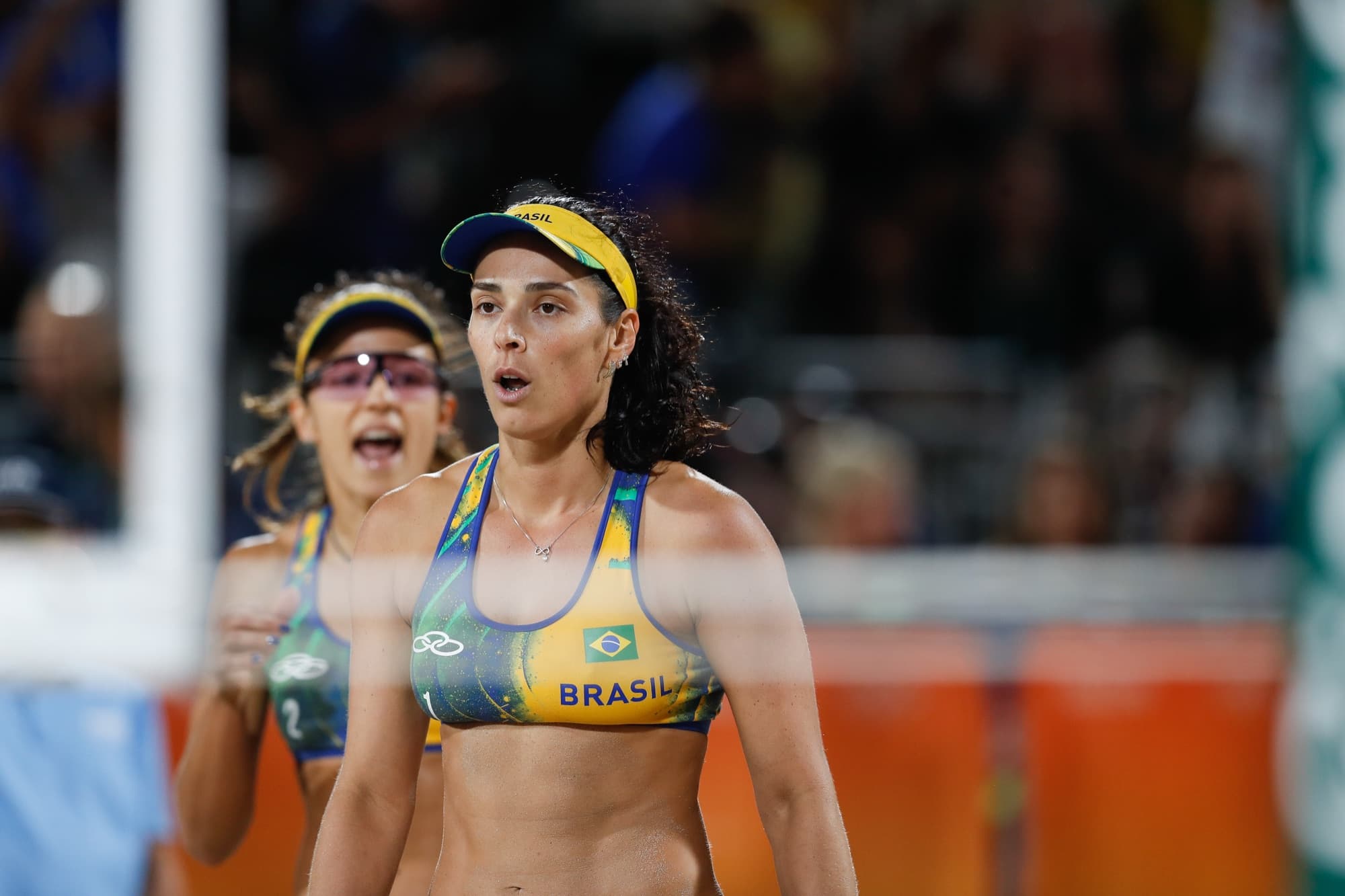 Dupla brasileira de vôlei de praia concentrada na areia durante a final, com a torcida ao fundo